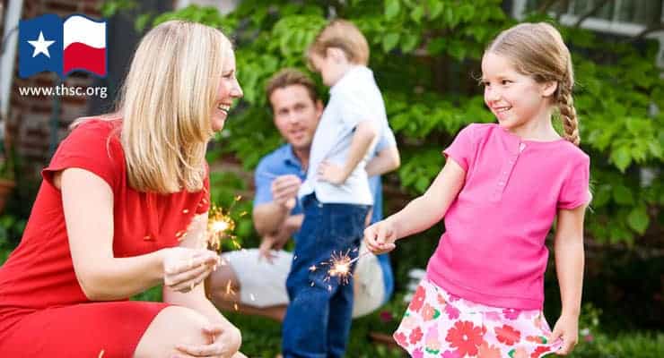 Mom teaches girl to hold sparklers, father, son in background
